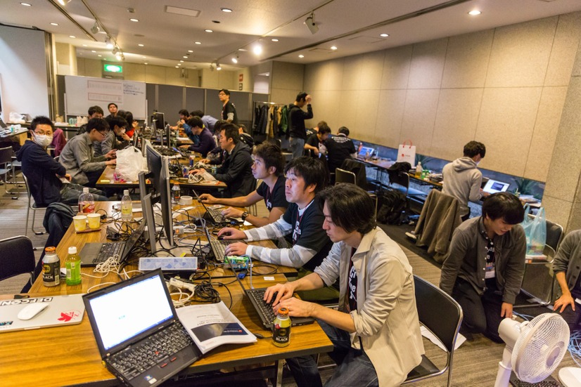 昨年開催された Trend Micro CTF 2017 の決勝戦を迎えた際のオペレーションセンター写真。手前のテーブルがインフラチーム、その奥のテーブルはスコアサーバ＆可視化サーバチーム、そのさらに奥のテーブルと写真右側で壁に向かうのは問題チーム（写真右下の扇風機は空調に問題があった場合の対策で持ち込まれた）